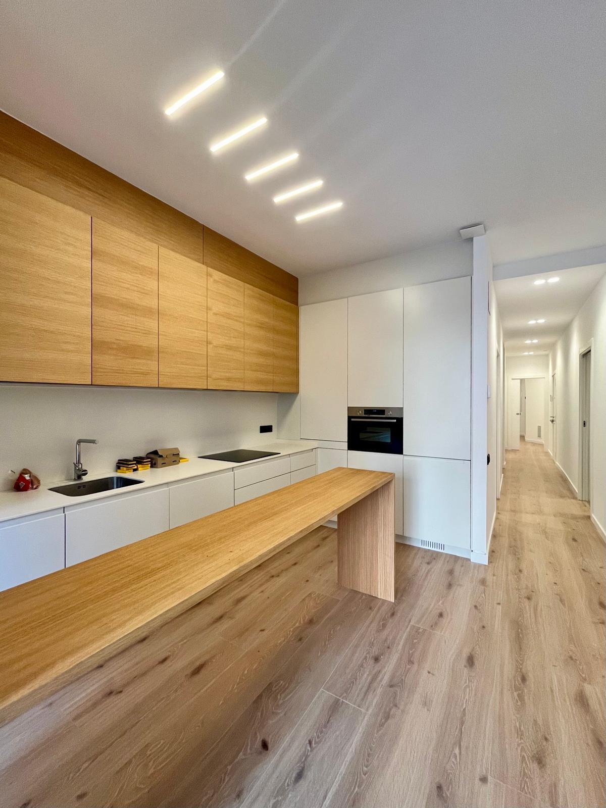 Cocina abierta con barra de roble, frentes blancos y el pasillo de la vivienda al fondo